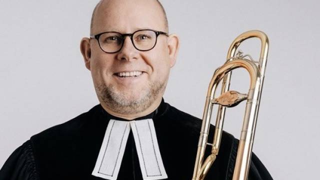 Porträt eines lächelnden Mannes in schwarzer Amtsrobe mit predigtstuhl-Krause, der eine Posaune in der Hand hält.