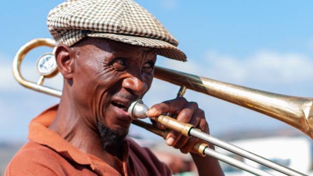 Ein Mann spielt im warmen Sonnenlicht Posaune. Er trägt ein orangefarbenes T-Shirt und eine Golfmütze. Die Szene wirkt hell und sommerlich.