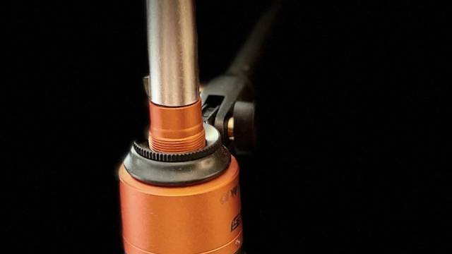 Ein einzelnes Mikrofon mit schwarzem Windschutz und orangefarbenem Metallgehäuse ist vor tiefschwarzem Hintergrund zu sehen. Das Kabel führt nach oben aus dem Bild. Die Beleuchtung hebt die Konturen des Mikrofons klar hervor.