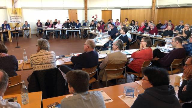 Viele Menschen sitzen in einem großen Tagungsraum an Tischen und hören einer Sitzung oder Präsentation zu. Auf den Tischen liegen Unterlagen, Getränke und Arbeitsmaterialien. Im Hintergrund hängt ein Banner des Evangelischen Posaunendienstes in Deutschland.