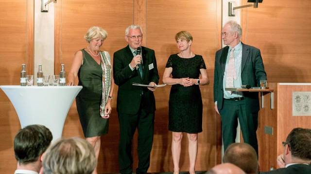 Vier Personen stehen bei einer offiziellen Veranstaltung auf einer Bühne, eine Person spricht in ein Mikrofon und hält Unterlagen in der Hand. Die anderen hören aufmerksam zu, auf Stehtischen stehen Wasserflaschen und Gläser. Die Szene wirkt feierlich und formell.