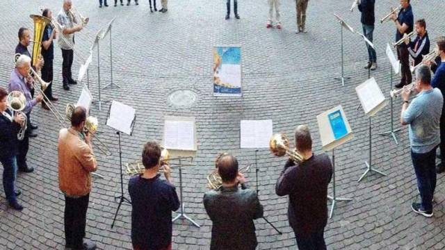 Ein Bläserkreis steht in einem weiten Kreis auf einem gepflasterten Platz und spielt nach Notenständern. In der Mitte liegt ein Plakat auf dem Boden. Die Musiker sind von oben fotografiert und wirken konzentriert beim gemeinsamen Musizieren.