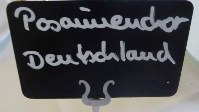 Ein kleines schwarzes Schild steht auf einem Metallhalter. Darauf ist mit weißer Handschrift „Posaunenchor Deutschland“ geschrieben. Das Schild wirkt schlicht und handgemacht.