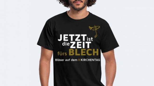 Zu sehen ist eine Person im T-Shirt mit gold-weißem Text: „JETZT ist die ZEIT fürs BLECH – Bläser auf dem #KIRCHENTAG“.