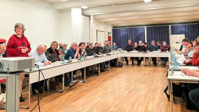 In einem großen Tagungsraum sitzen zahlreiche Personen an U-förmig angeordneten Tischen und verfolgen eine Sitzung. Auf den Tischen stehen Wasserflaschen, Unterlagen und technische Geräte. Eine Person im roten Pullover steht links und spricht in die Runde.