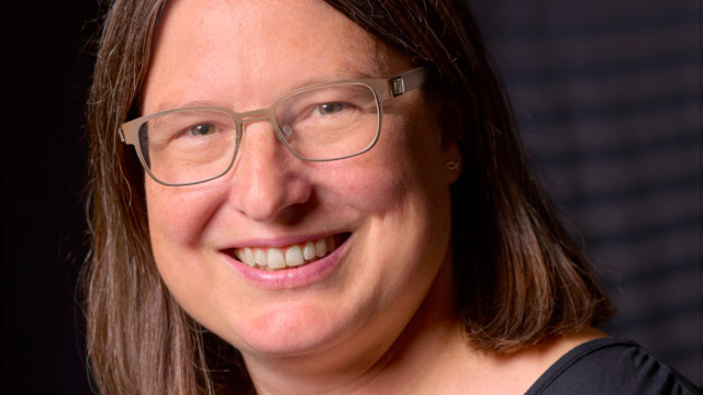 orträt einer lächelnden Frau mit schulterlangem braunem Haar und Brille vor dunklem Hintergrund