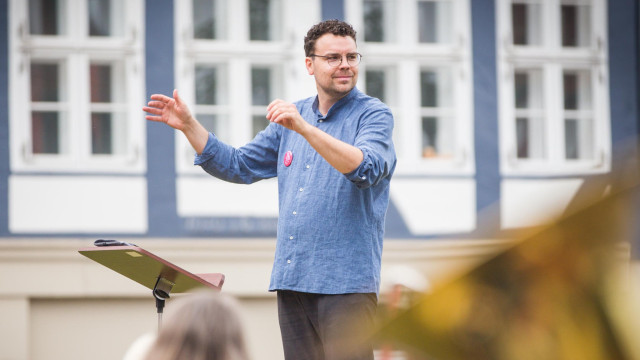 Dirigent mit Brille leitet im Freien vor Gebäude ein Ensemble.