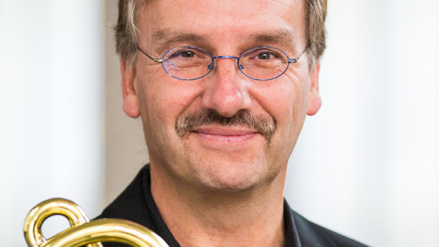 Porträt eines lächelnden Mannes mit Schnurrbart und ovaler Brille; im Vordergrund ist ein goldenes Blechblasinstrument zu sehen, Hintergrund hell und unscharf