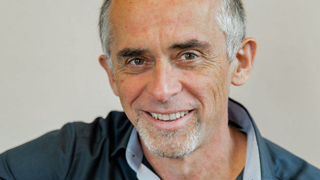 Nahporträt eines lächelnden Mannes mit stark zurückgehendem grauem Haar und hellem Stoppelbart am Kinn; er trägt ein dunkelblaues Poloshirt mit aufgeklapptem Kragen, darunter ein weißes Shirt, Hintergrund: einfarbige helle Wand